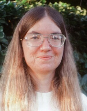 Carol Shaw holding her gold River Raid cartridge (cropped) / Portrait of Carol Shaw wearing glasses, a white top, and with long fair hair. This image was cropped and uploaded to Wikipedia. / Image credit: Carol B Shaw / This work is licensed under Creative Commons Attribution-ShareAlike 4.0