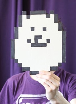 Cropped photo from a 2017 Famitsu interview with Toby Fox / A portrait of Toby Fox obscuring his face with a painted paper mask of his personal avatar, the character Annoying Dog (a small white Pomeranian that first appeared in Undertale). / Image credit: Famitsu